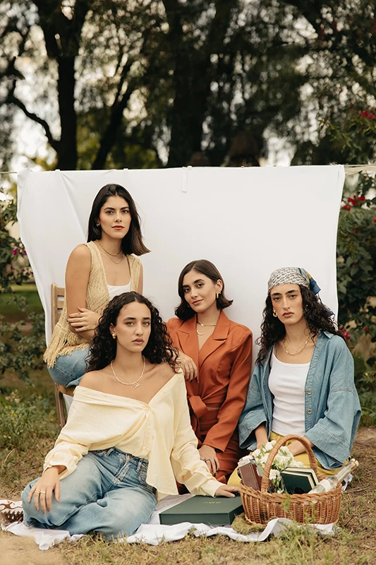 Cuatro modelos femeninas sentadas sobre el cesped con fondo de una tela blanca. Todas lucen productos exclusivos de Mena Fine Jewelry.