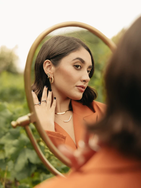 Modelo femenina mirandose a si misma en un espejo, mientras luce collares y aretes de Mena Fine Jewerly.