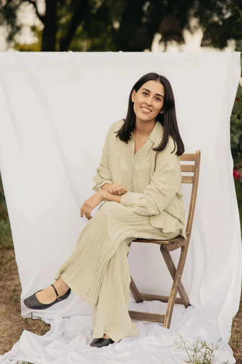 Ximena, creadora y diseñadora de Mena Fine Jewelry, sentada con pose relajada y piernas cruzadas en una silla de madera con un fondo de tela blanco.