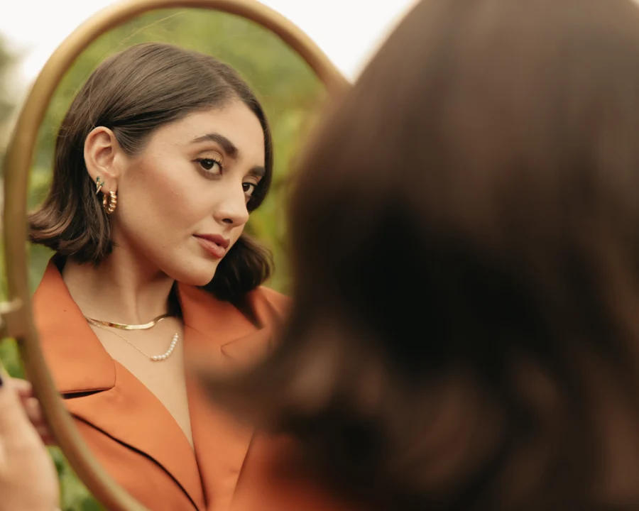 Modelo femenina mirandose a si misma en un espejo, mientras luce collares y aretes de Mena Fine Jewerly.