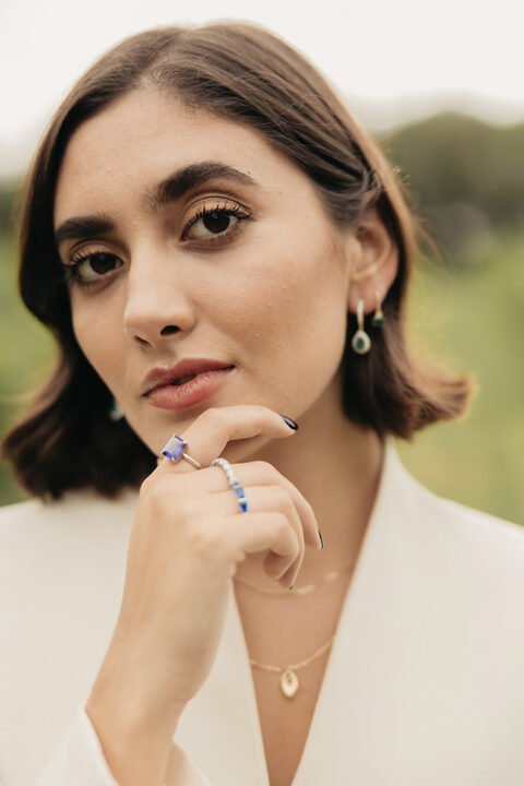 Primer plano de una modelo con una mano en su mentón, luciendo aretes, collares y anillos con piedras. Diseños originales de Mena Fine Jewelry.